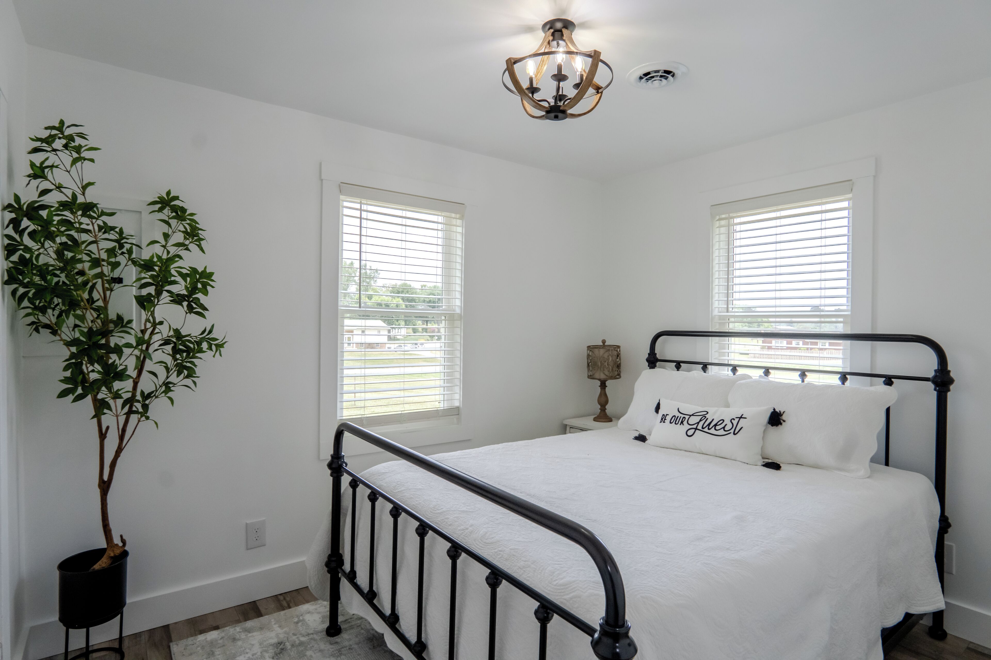 A bedroom with a bed, two windows, a chandelier, a bedside table, and a potted plant.