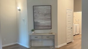 A hallway with wooden flooring, a white console table with drawers, a wall-mounted abstract painting, a white door, and a small potted plant on the console table.