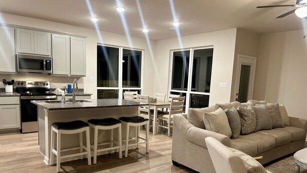 A modern kitchen with a central island, bar stools, a dining area with a table and chairs, and a living room with a sofa and armchair.