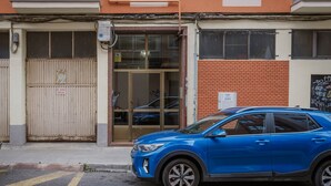 A blue car parked in front of a building with a beige gate, a glass door, and a brick wall.