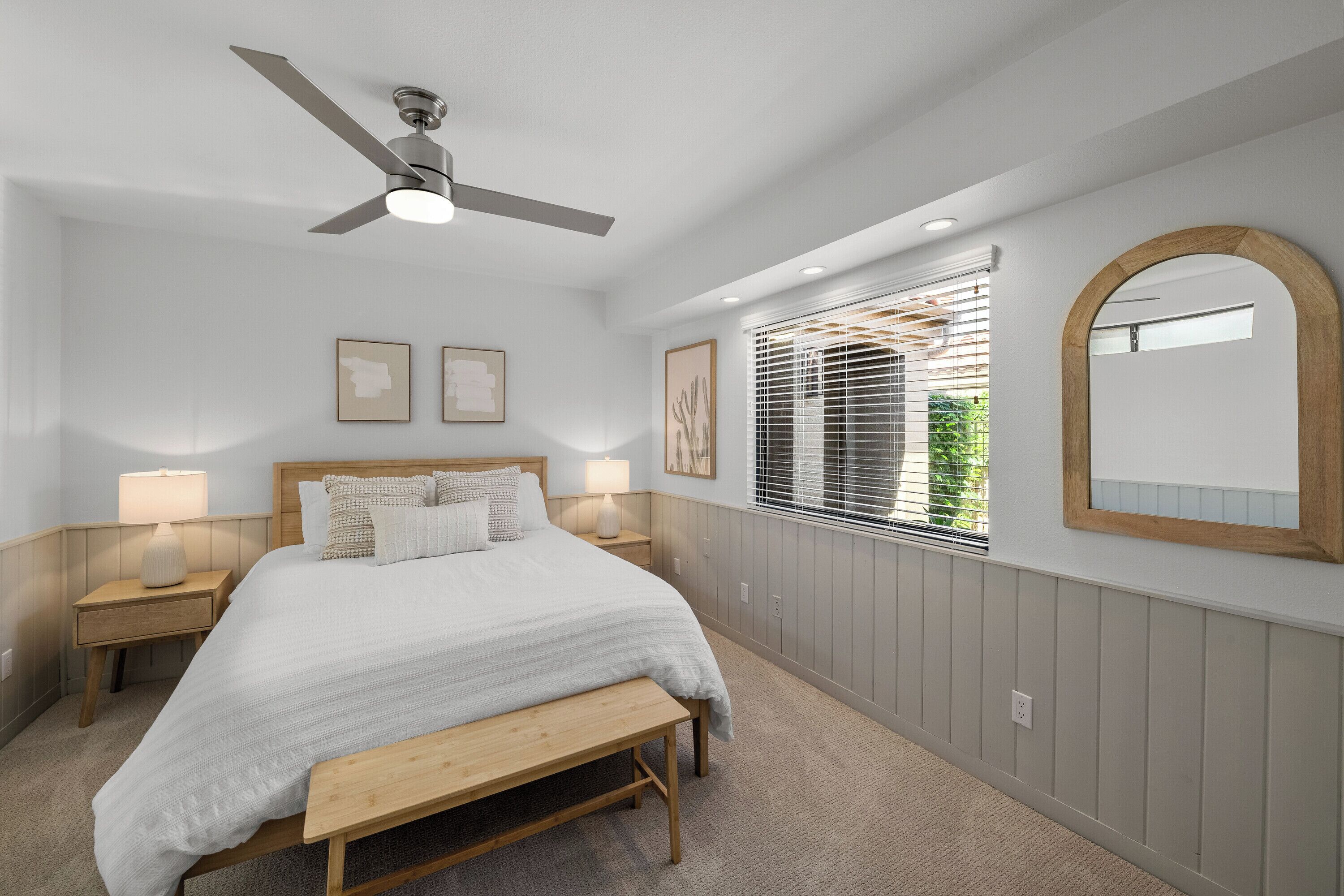 A bedroom with a bed, bedside tables, a ceiling fan, a window with blinds, and a mirror on the wall.
