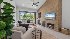 A modern living room with a fireplace, a large window, and a ceiling fan. - 3 Bedroom Luxury Condo on Fairway at Palm Valley CC! Available January 2027! (Palm Desert)