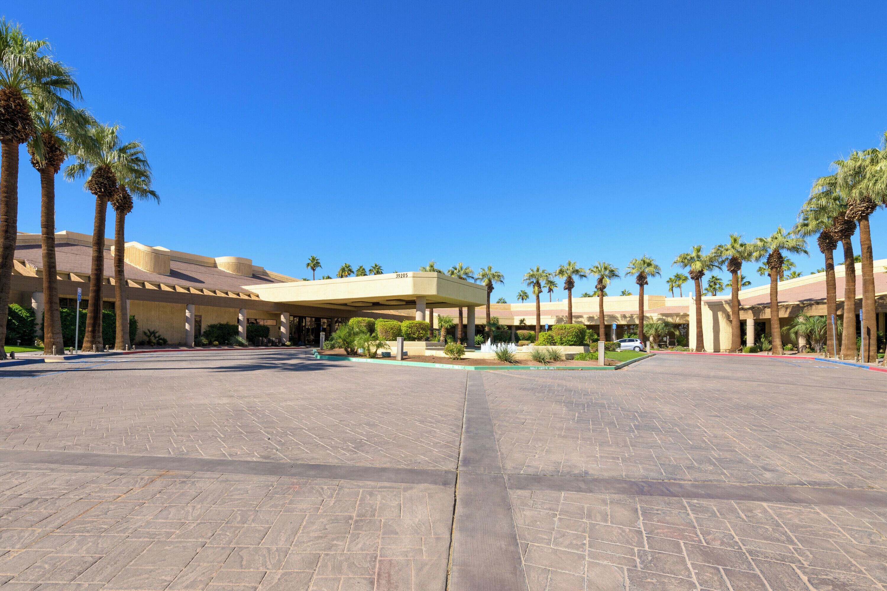 A spacious courtyard with a large building featuring a flat roof and columns, surrounded by palm trees.
