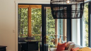 A modern interior with a dining area, a sofa, and a large window with a view of greenery. - Cabine Origine  (Lac-Supérieur)