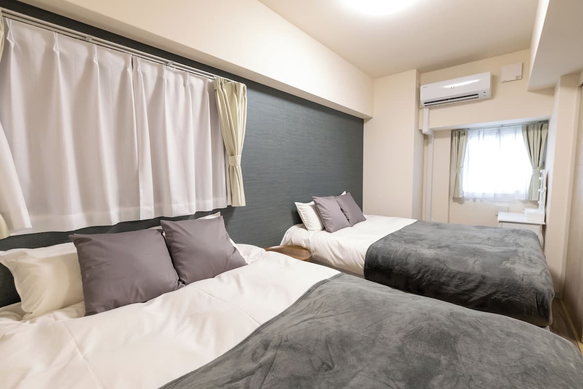 Une chambre d’hôtel avec deux lits, une tête de lit grise, une literie blanche et grise, une fenêtre avec des rideaux et un appareil de climatisation.