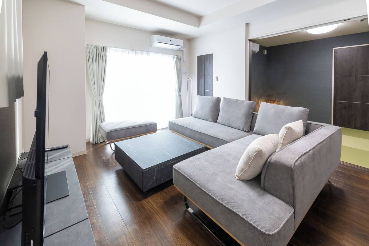 Un salon moderne avec un canapé gris, un téléviseur à écran plat et une grande fenêtre agrémentée de rideaux.
