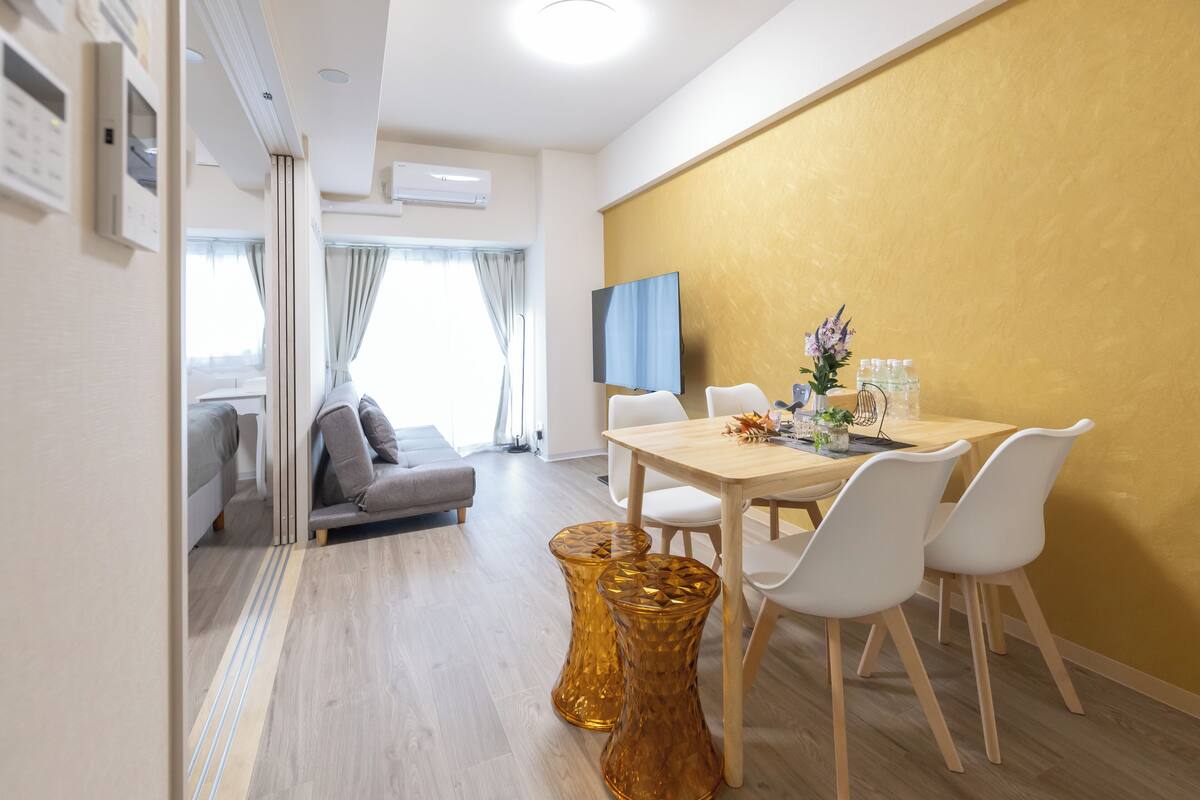 Un salon moderne avec un canapé, une table à manger et des chaises, un téléviseur et un mur d’accent jaune.