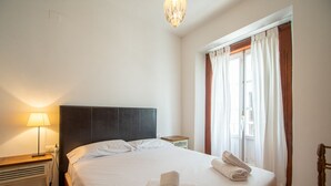 Ein ordentlich bezogenes Bett mit weißen Laken und Handtüchern, eine Nachttischlampe und ein Fenster mit Vorhängen.