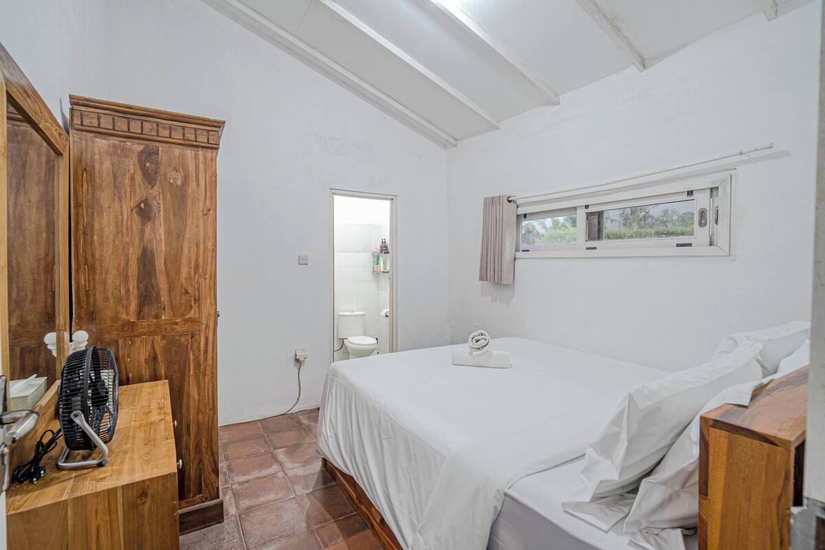 Een slaapkamer met een houten kledingkast, een bed met witte lakens, een raam met gordijnen en een ventilator op een houten tafel.