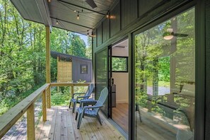 Eine Holzterrasse mit Adirondack-Stühlen, eine Glastür, die zu einem Raum führt, und Blick auf einen Wald.