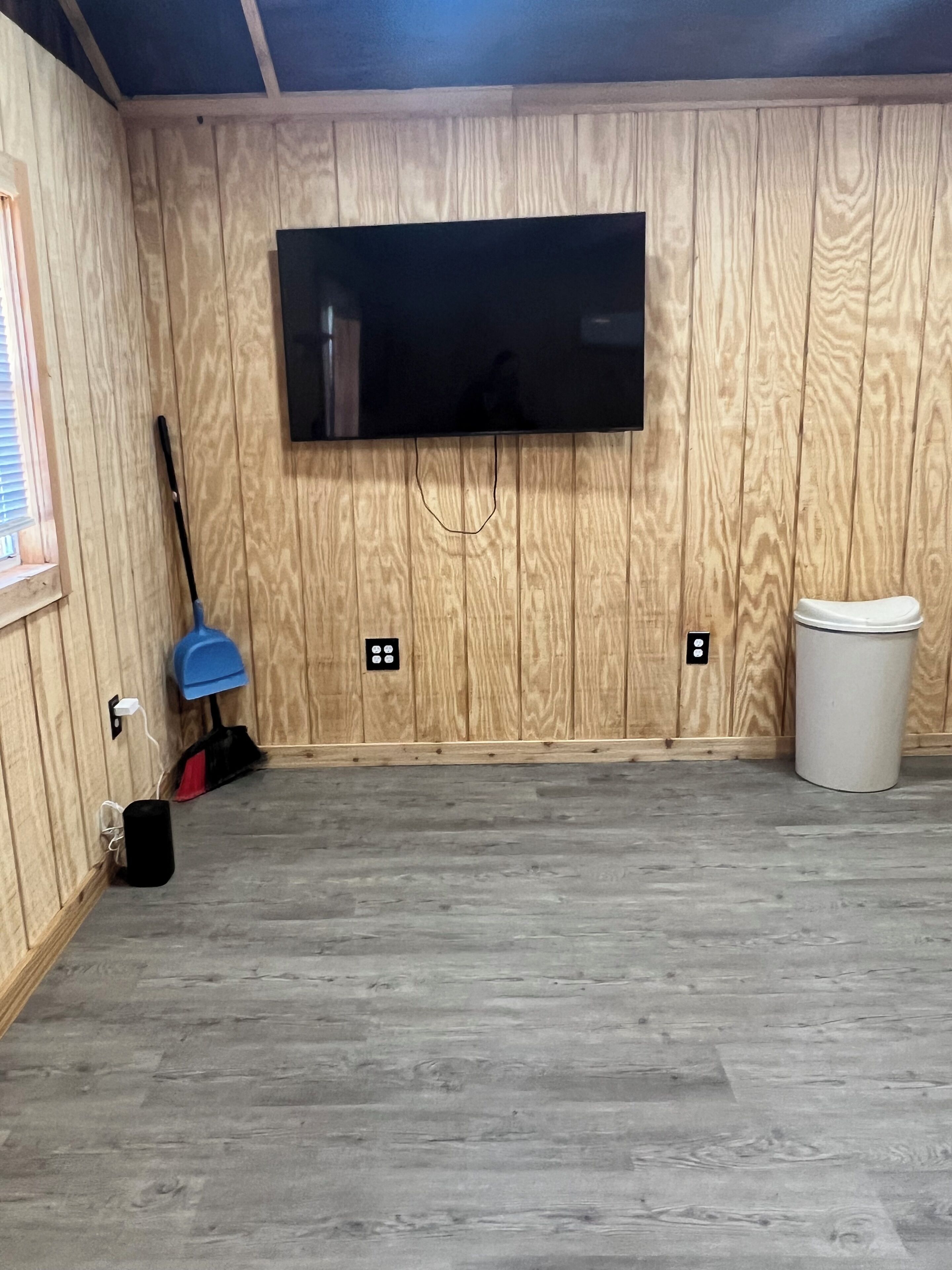 Habitación con paredes de paneles de madera, televisor montado, escoba, cubo de basura y toma de corriente.