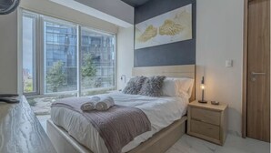 Ein Schlafzimmer mit einem großen Fenster, einem Bett mit zusammengerollten Handtüchern, einem Nachttisch und einem Wandbild.