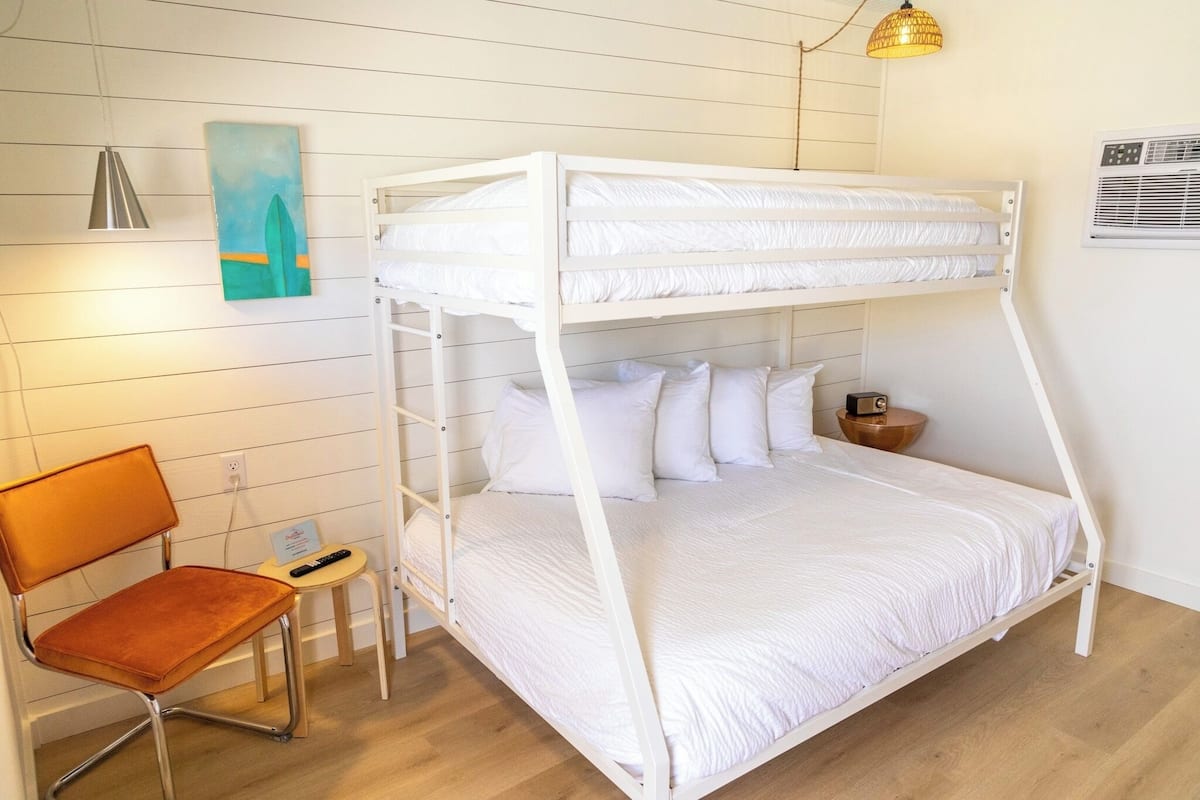 A bunk bed with white bedding, a wooden chair, a small table, and a painting on the wall.