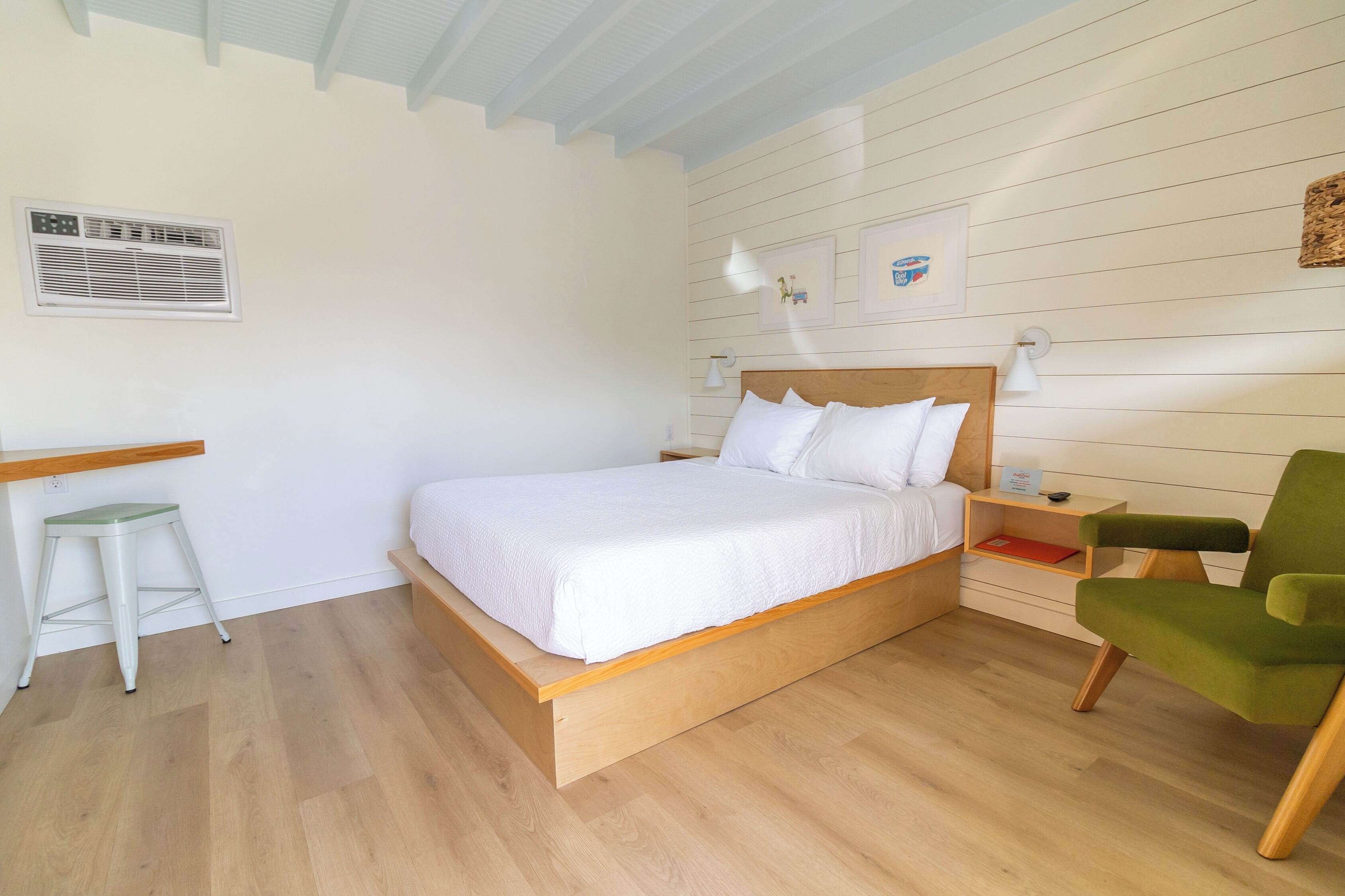 Un dormitorio con una cama de madera, una silla verde, un taburete blanco, una mesita de noche y una lámpara de pared.