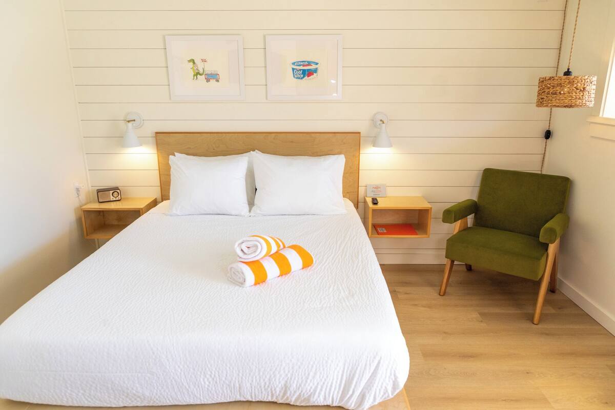 A neatly made bed with white linens and two pillows, a green armchair, and two bedside tables with lamps and a radio.