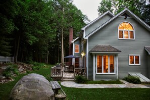 A two-story house with a deck, a chimney, and a large rock in the yard.