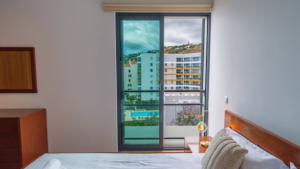 Une chambre d’hôtel avec un lit, une table de chevet, une lampe et une vue sur un immeuble et une piscine, visibles à travers une porte vitrée.