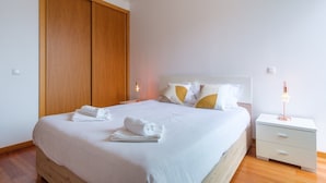 Un lit bien fait, avec du linge de lit et des serviettes blanches, une armoire en bois, une table de chevet avec une lampe et une commode blanche.