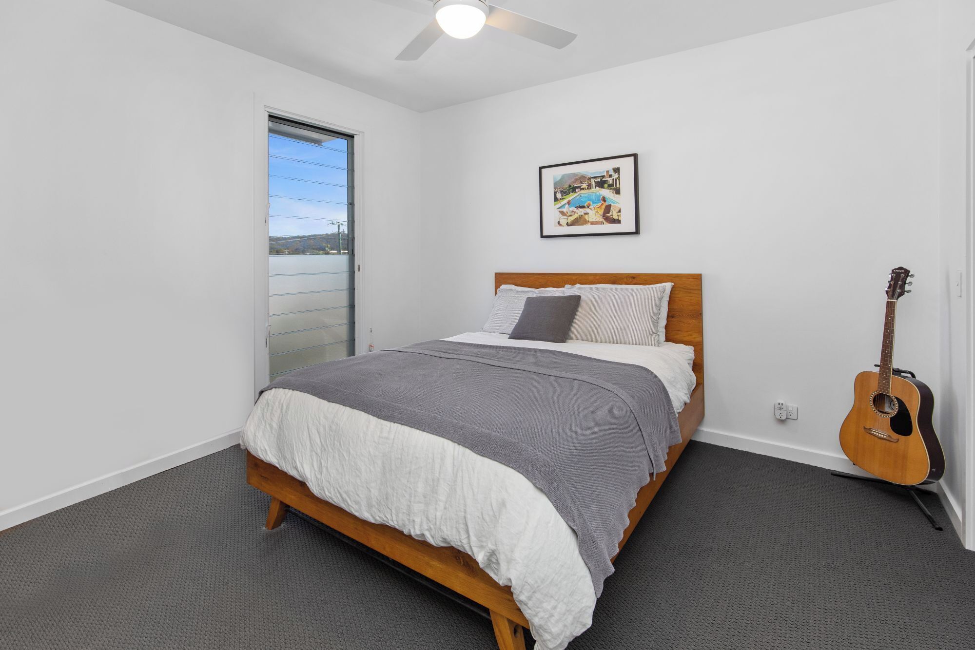 A bedroom with a bed, a guitar, a painting, and a view of a body of water through a sliding door.