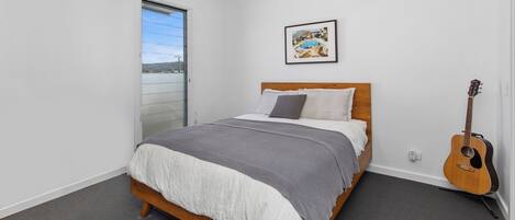 A bedroom with a bed, a guitar, a painting, and a view of a body of water through a sliding door.