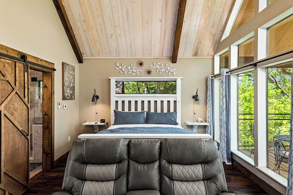 Un dormitorio con una cama grande, vigas de madera en el techo y vistas al exterior con zonas verdes, además de una puerta corrediza a la izquierda.