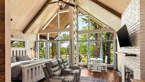 A cozy room with a bed, a reclining chair, a fireplace, and large windows offering a view of greenery. - Fairway Treehouses (Berryville)