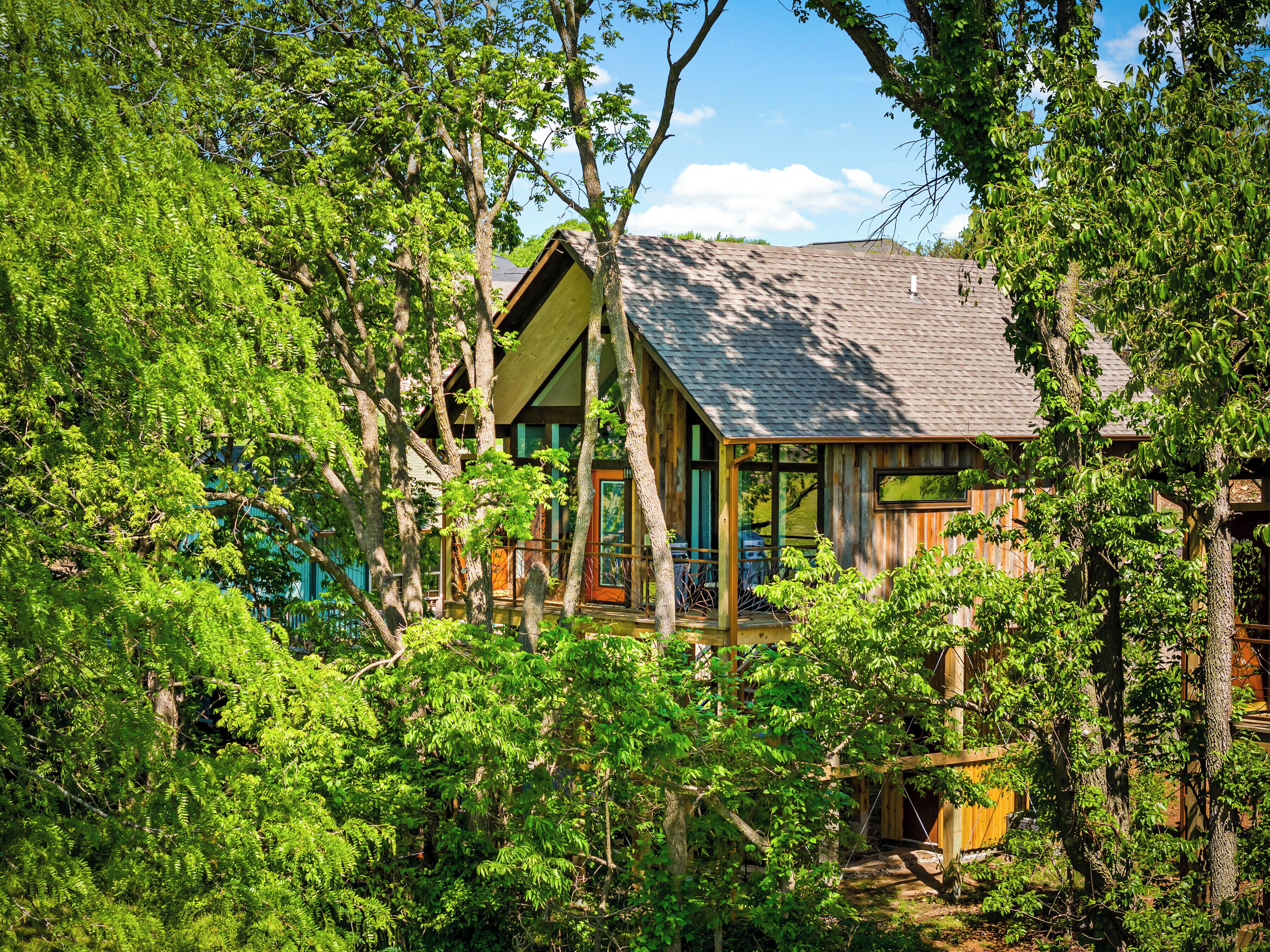 Un chalet rustique entouré d’une forêt dense, doté d’une véranda et de grandes fenêtres.