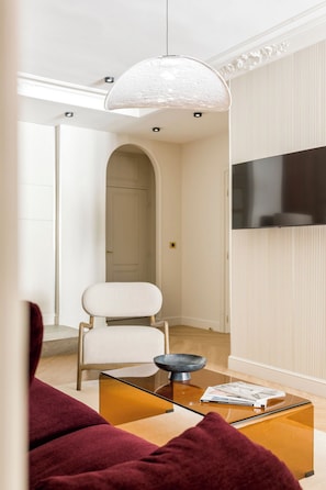 A modern living room with a glass coffee table, a white armchair, and a large TV mounted on the wall. - Merveil - Signature Suite -Tour Eiffel- Grenelle I (Paris)