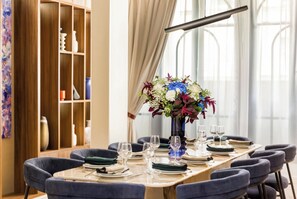 A dining area with a long table set for six, surrounded by chairs. A vase with flowers is placed on the table, and there is a bookshelf with various items on the left. - Merveil - Signature Suite -Tour Eiffel- Grenelle I (Paris)