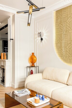 A modern living room with a sofa, a coffee table with magazines and a glass, a decorative lamp, and a patterned wall. - Merveil - Signature Suite -Tour Eiffel-Grenelle II (Paris)
