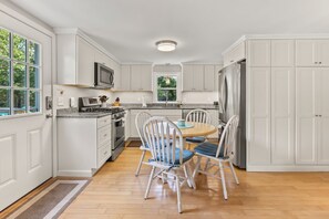 A kitchen with white cabinetry, a stainless steel refrigerator, a microwave, and a gas stove. There is a dining table with chairs and a window with a view of greenery outside. - Cape Cod Cottage Near Beaches & Bike Trail (Harwich)