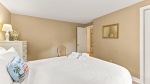 A hotel room with a large bed, a nightstand with a lamp, a chair, and a door leading to another room. - Cape Cod Cottage Near Beaches & Bike Trail (Harwich)