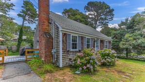A single-story house with a brick chimney, a covered deck, and a garden with hydrangea bushes. - Cape Cod Cottage Near Beaches & Bike Trail (Harwich)