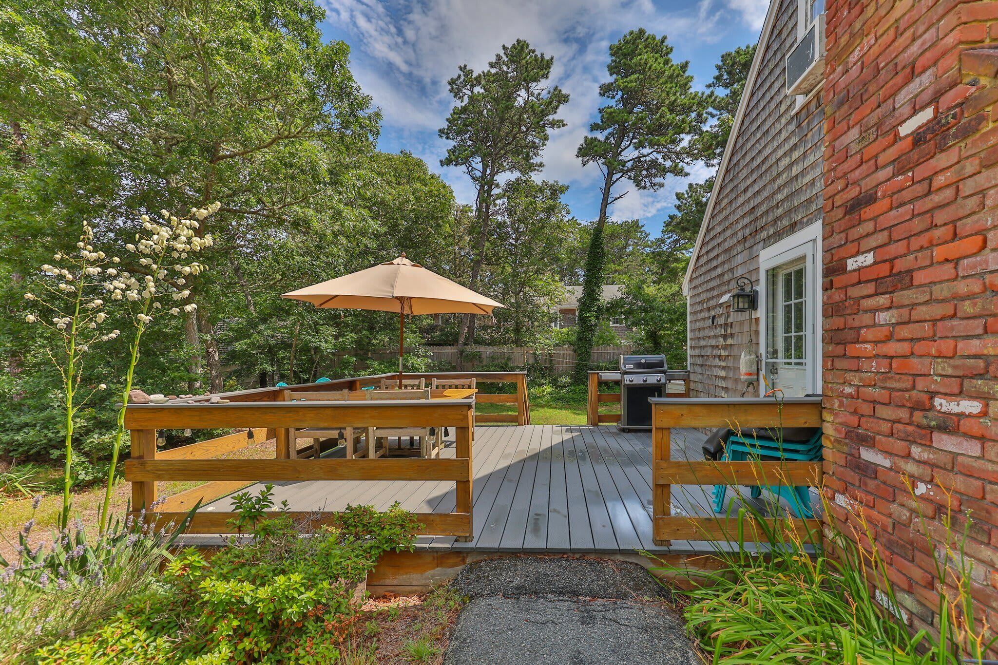 A wooden deck with benches and a barbecue grill, surrounded by greenery and a brick building.