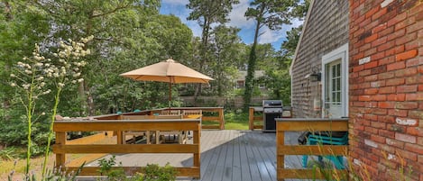 A wooden deck with benches and a barbecue grill, surrounded by greenery and a brick building.