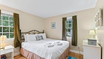 A neatly made bed with white linens and a pink skirt, a bedside table with a lamp, and a blue and white striped rug on a wooden floor.