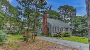 A rustic cabin with a chimney, surrounded by trees and a wooden deck. - Cape Cod Cottage Near Beaches & Bike Trail (Harwich)