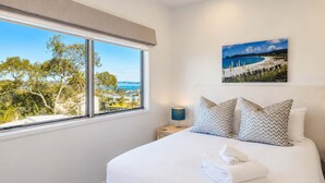 A bedroom with a large window, a bed with white linens, two pillows, a bedside lamp, and a painting of a beach scene.