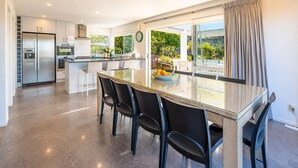 A modern kitchen with a central island, a dining area with a glass table, and a view of greenery outside.