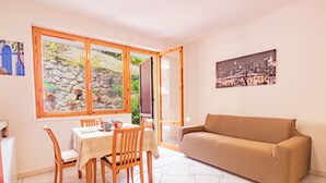 A cozy room with a dining table, chairs, a sofa, and a large window with a view of a stone wall and greenery. - Twin House - Le Lecce (Capoliveri)