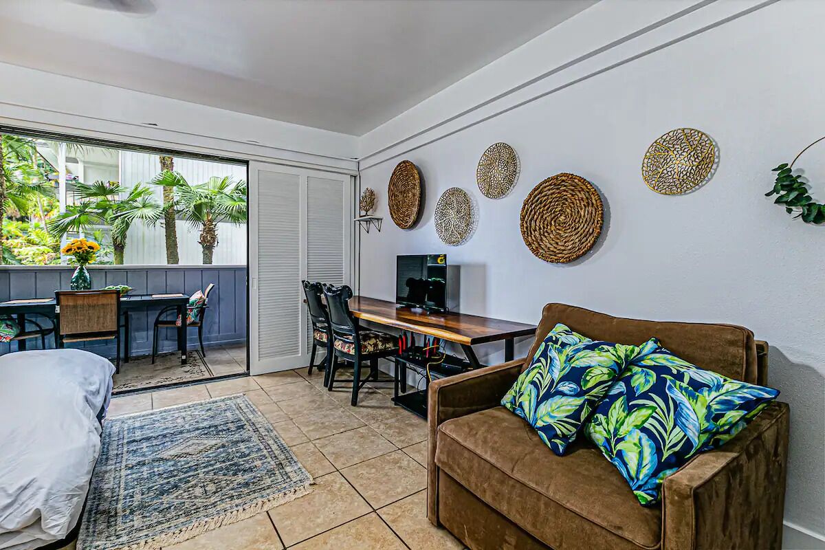 A modern living room with a sofa, a dining table, and a TV. There is a balcony with plants and a view of palm trees.