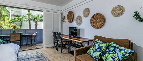 A modern living room with a sofa, a dining table, and a TV. There is a balcony with plants and a view of palm trees.