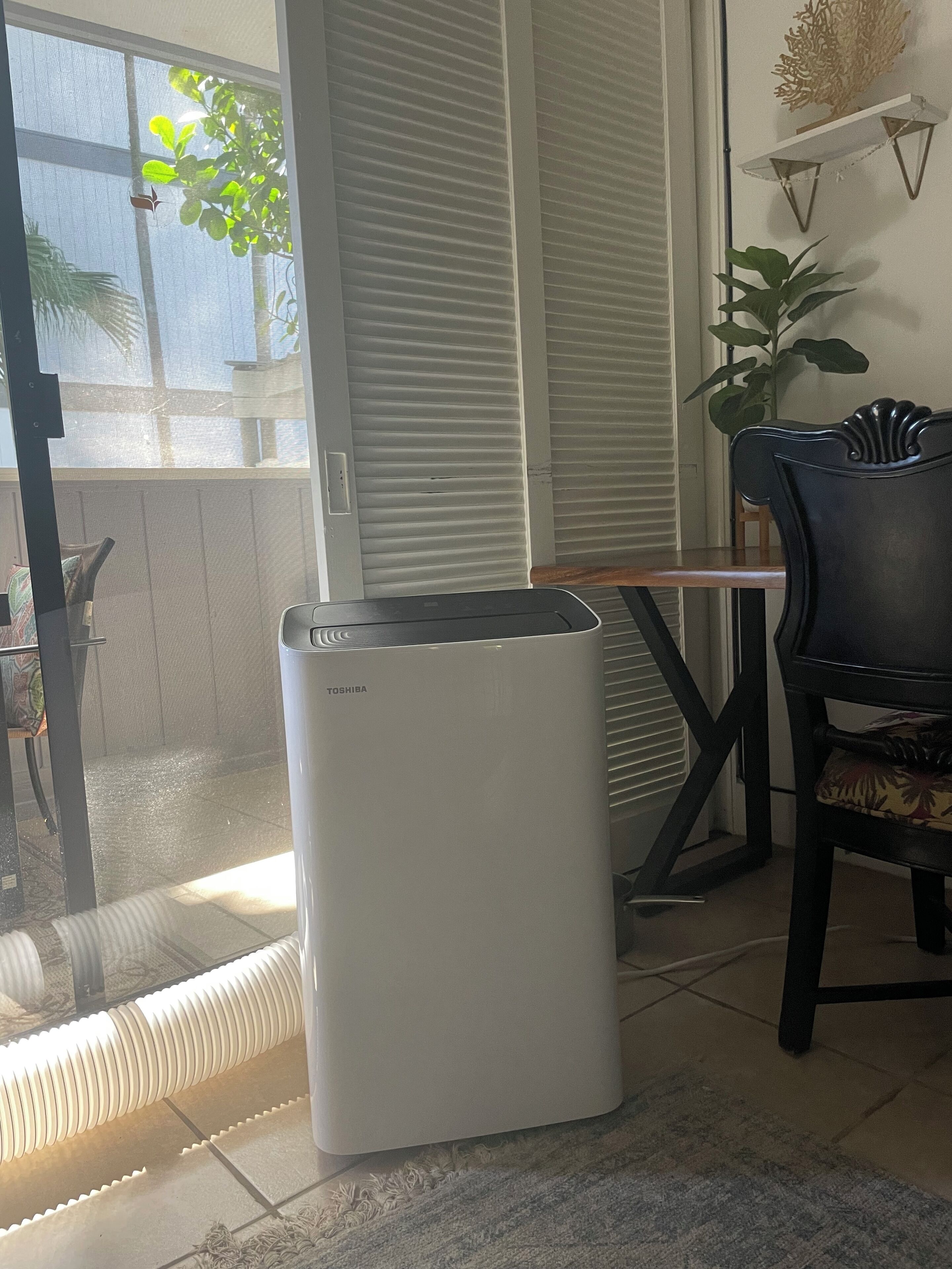 A white air conditioner in a room with a sliding glass door, a wooden table, and a black chair.