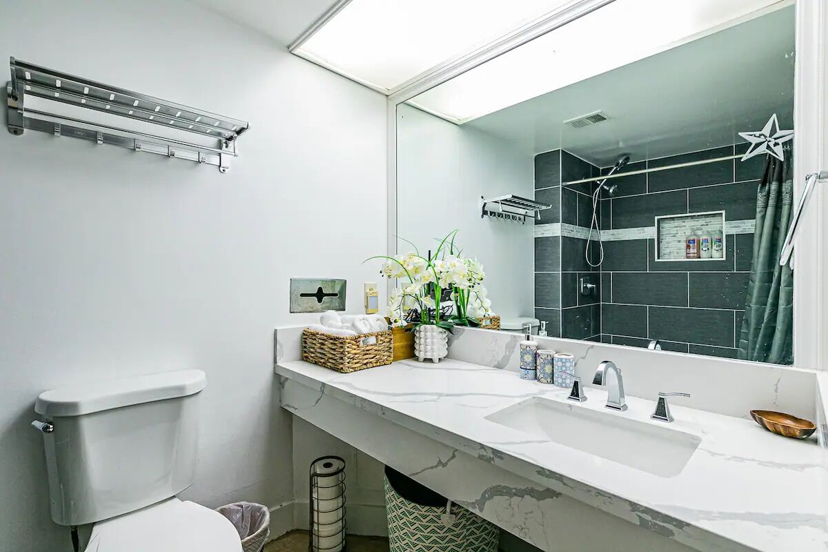 A modern bathroom with a white toilet, a large mirror, a sink with two faucets, and a shower area.