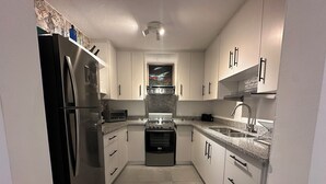 Cocina compacta con armarios blancos, refrigerador de acero inoxidable, horno empotrado y encimera de granito.