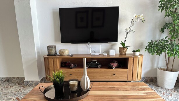 Una mesa de madera con una bandeja de objetos, un televisor montado en la pared y un mueble de madera decorado con adornos y plantas.