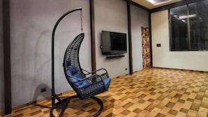 A spacious room with a wooden parquet floor, a hanging chair, a flat-screen TV mounted on the wall, and a large window. - Wandr Tryo Villa Karjat (Khalapur)