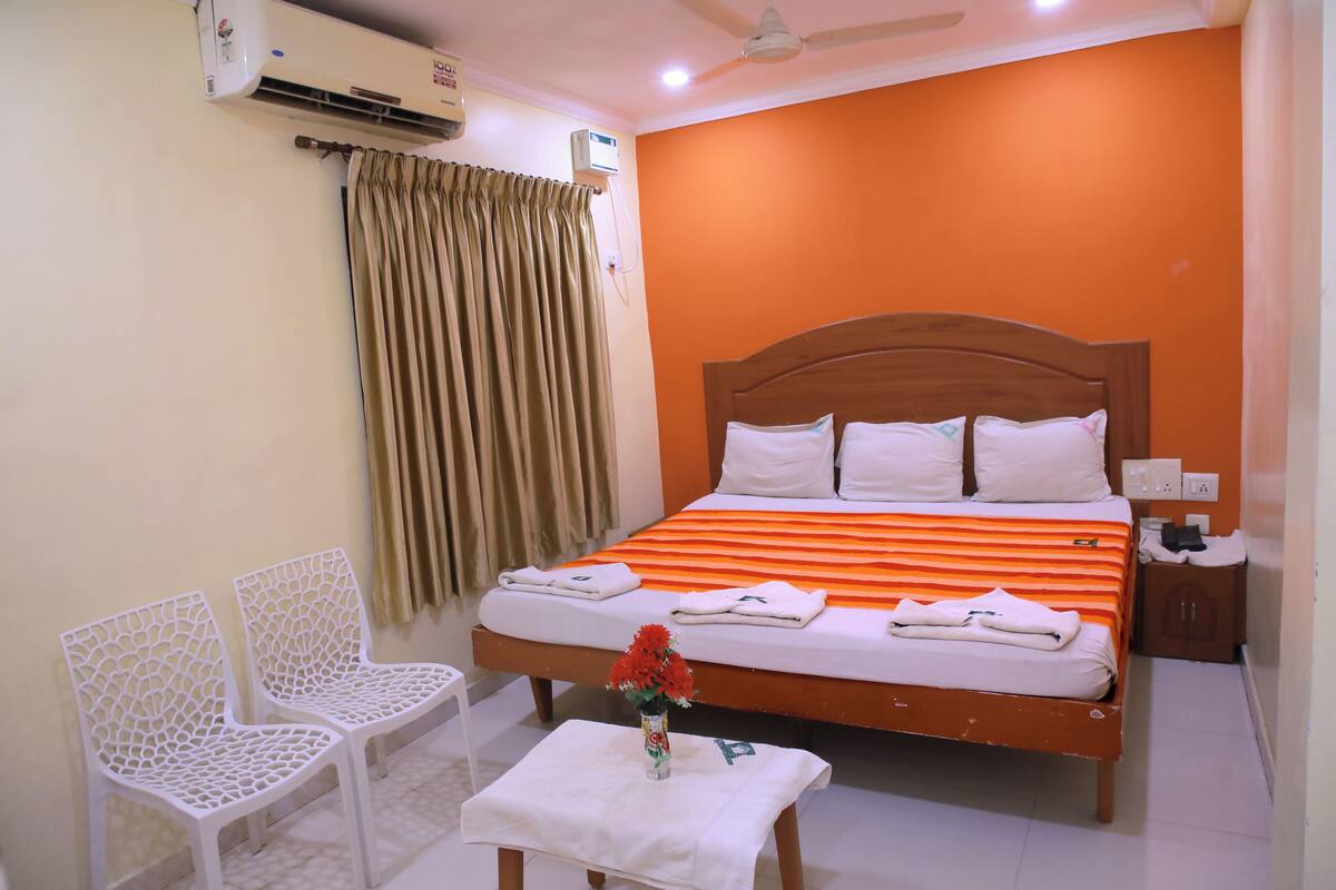 A hotel room with a bed, two white chairs, a small table, an air conditioner, and a ceiling fan.