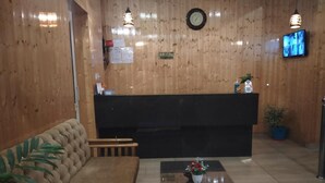 Un'area di accoglienza con parete in legno, scrivania della reception, un divano, un tavolino con un vaso di fiori e un televisore a parete.