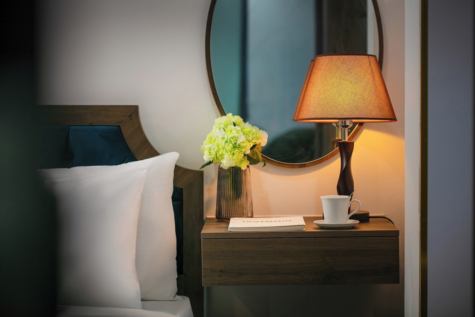 A neatly made bed with white linens, a bedside table with a lamp, a vase with flowers, and a cup and saucer.